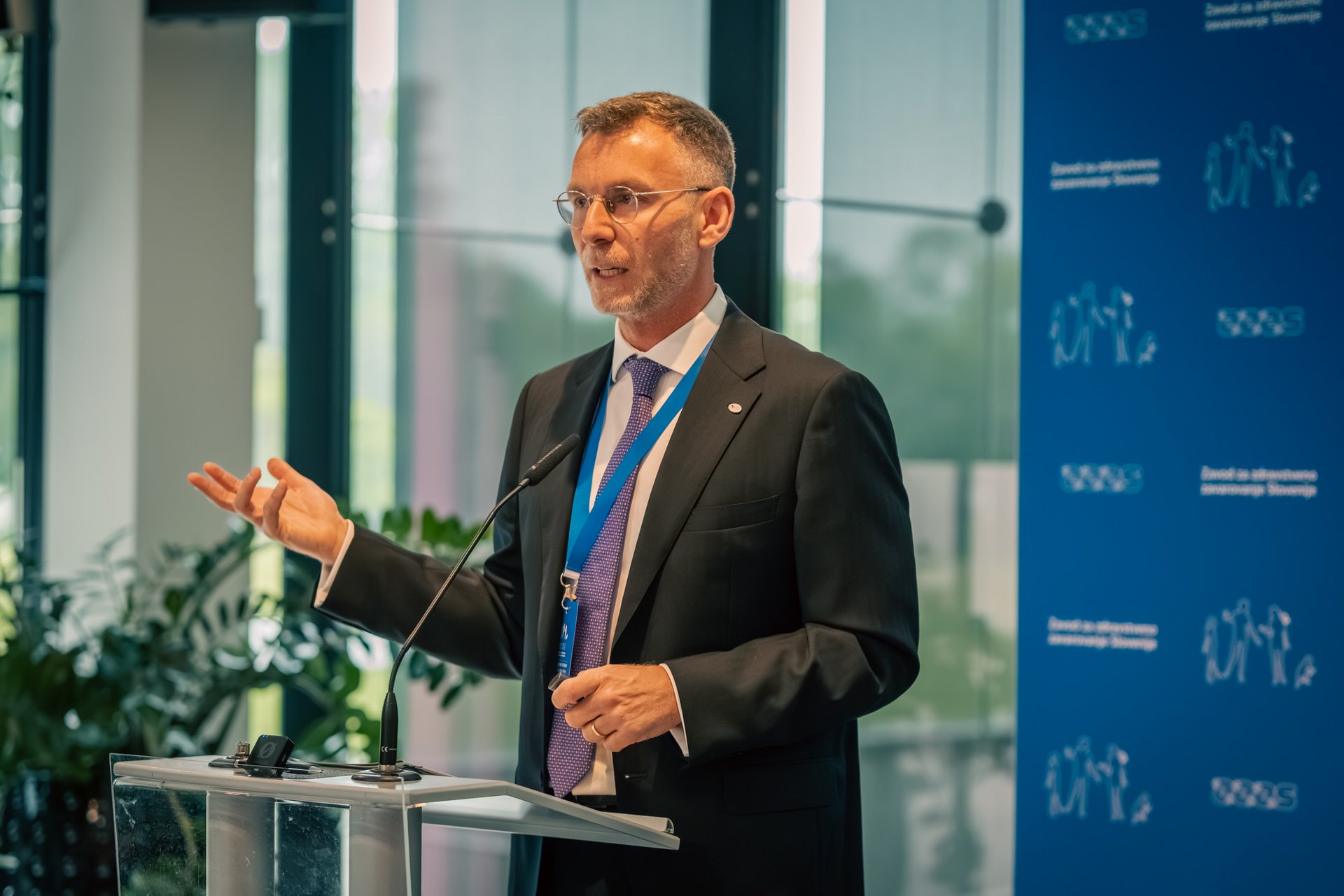Yannis Natsis, direktor Evropskega združenja nosilcev obveznih socialnih zavarovanj (ESIP)2, za govorniškim pultom, zadaj modra stena z logotipi ZZZS.