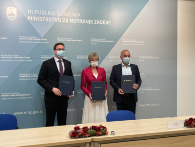 Tri osebe (Boštjan Koritnik, Tatjana Mlakar, Aleš Hojs) stojijo pred panojem ministrstva za notranje zadeve in držijo v rokah posebne mape; vsi nosijo zaščitne maske
