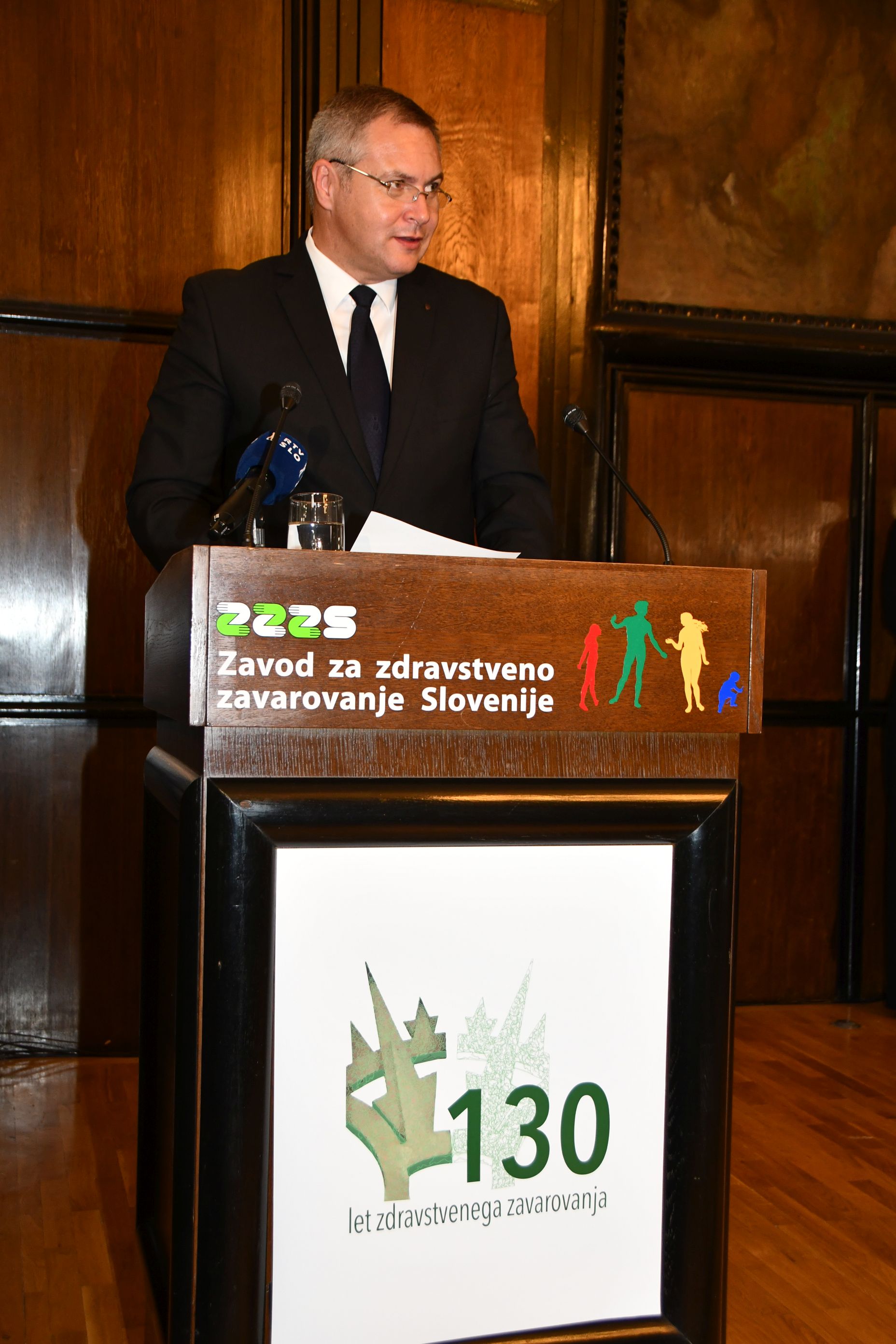 Gospod (Dejan Židan, predsednik Državnega zbora RS) v temni obleki s kravato in očali stoji za lesenim govorniškim odrom z napisom ZZZS in 130 let in govori