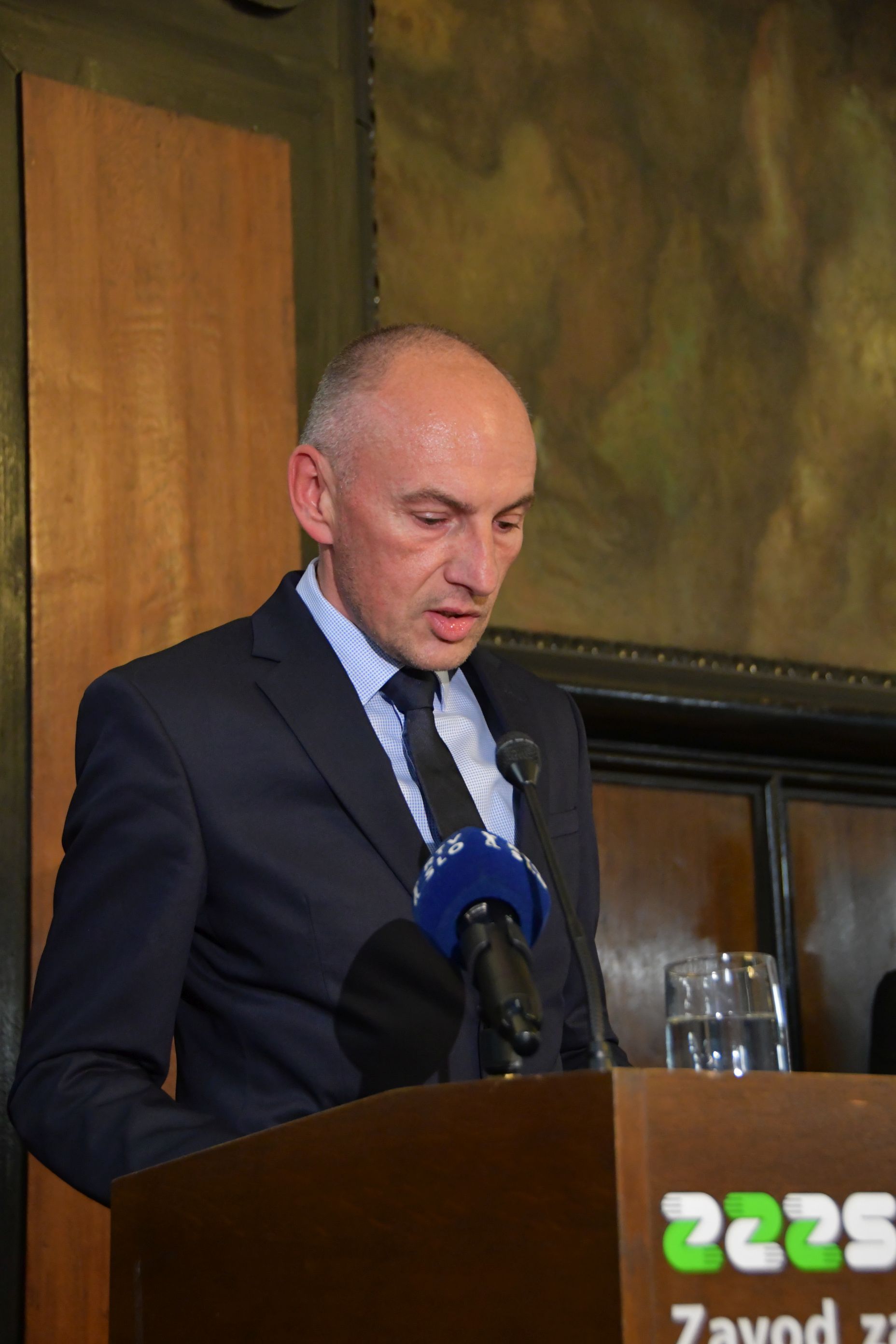Gospod (Aleš Šabeder) v temni obleki s kravato stoji za lesenim govorniškim odrom z napisom ZZZS in govori